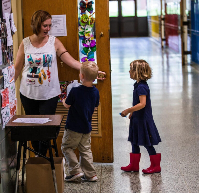 Two children entering a classroom greeted by female teacher
